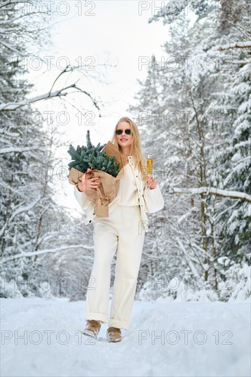 Low angle view of young cheerful woman standing on snowy forest road with fir tree branches and champagne
