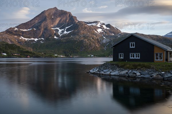 Landscape on the Lofoten Islands. A wooden house in Kakern on Kakersundet. The mountain Narvtinden on Moskenesoya in the background. The mountain and the house are reflected in the sea. At night at the time of the midnight sun in good weather, a few clouds in the sky. Early summer. Kakern, Flakstadoya, Lofoten, Norway, Europe
