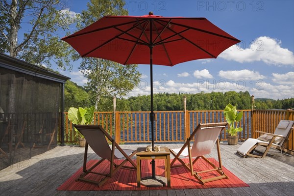 Black fishnet gazebo with red canvas cloth parasol and wooden folding canvas chairs on red mat on grey wood plank deck with railing overlooking lake with green forest in backyard in summer, Quebec, Canada, North America