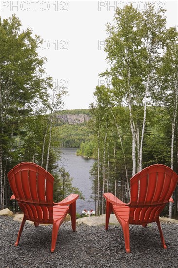 Two bright red plastic Adirondack chairs overlooking a calm lake through Betula, Birch trees with green leaves from gravel surface patio in backyard of a secluded log cabin home in late summer, Quebec, Canada, North America