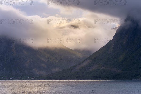 View over the Flakstadpollen to mountains in clouds with incoming sunlight. The small village of Flakstad in front. At night during the midnight sun. Early summer. Flakstadoya, Lofoten, Norway, Europe