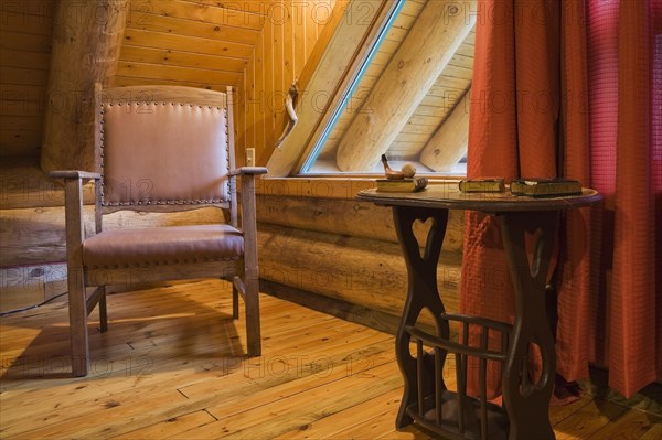 Brown wooden leather armchair and side table in master bedroom with red curtains and wood plank floor on upstairs floor inside rustic contemporary Scandinavian log cabin home, Quebec, Canada, North America