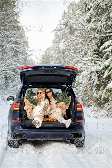 Two women are sitting in the trunk of a car holding in hands bouquets of fir branches and wine glasses