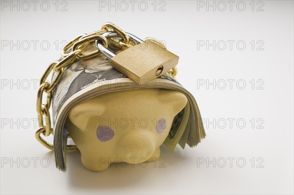 Close-up of wad of US currency bank notes wrapped with a brass chain and lock around a painted yellow piggy bank on a white background, Studio Composition, Quebec, Canada, North America