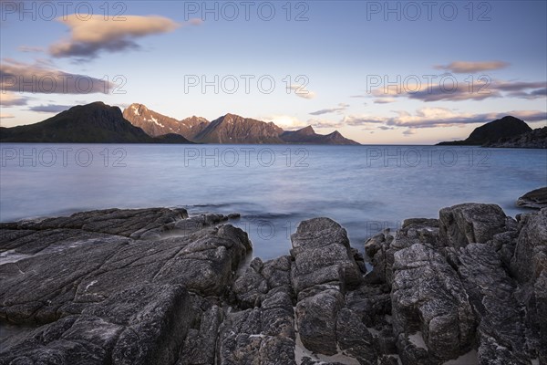 Seascape on the beach at Haukland. View of the mountains of Myrland on Flakstadoya. Rocks in the foreground. At night at the time of the midnight sun. Good weather, blue sky, a few coloured clouds. Early summer. Long exposure. Haukland Beach, Haukland, Vestvagoya, Lofoten, Norway, Europe
