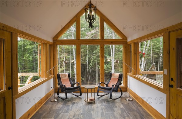 Reading room with tan and dark grey nuanced ceramic tile floor and two black leather rocking chairs plus pendant medieval candle light bulb style black wrought iron chandelier inside luxurious contemporary timber framed and milled log cabin home, Quebec, Canada, North America