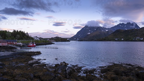 Landscape on the Lofoten Islands near Ramberg. On the left a few houses by the sea and a boat. Further back the Flakstad bridge (Flakstadbruene) and mountains on Moskenesoya. At night during the midnight sun. Good weather, some lenticular clouds in the sky. Early summer. Flakstadoya, Lofoten, Norway, Europe