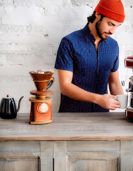 Focused barista in a blue patterned shirt and red beanie uses a manual coffee grinder, Vertical aspect ratio, AI generated