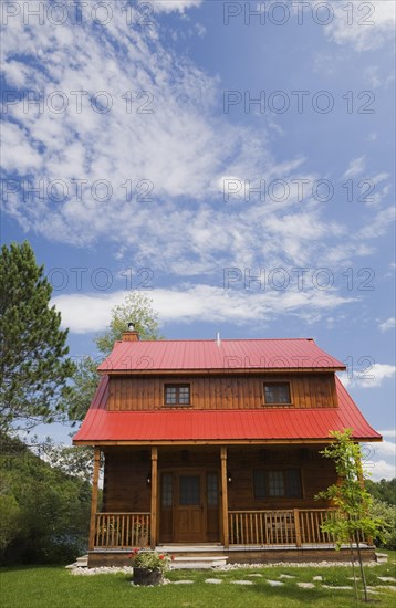 Small two story brown stained log cabin home with veranda and red sheet metal roof and landscaped front yard with deciduous tree and flagstone footpath in summer, Quebec, Canada, North America