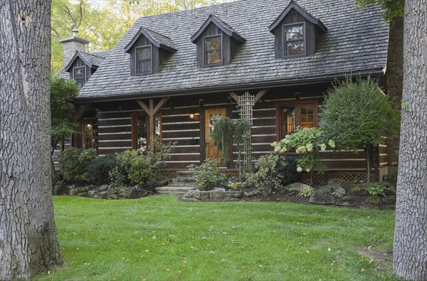 White chinked log cabin home facade with grey weathered cedar shingles roof, three dormer windows and landscaped front yard with mixed perennial shrubs and plants in borders in autumn, Quebec, Canada, North America