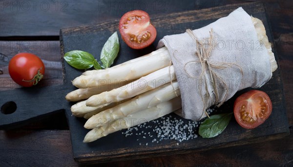 Fresh asparagus bundled on a wooden board next to tomatoes and basil, bunch of white asparagus wrapped in a damp kitchen towel, KI generated, AI generated