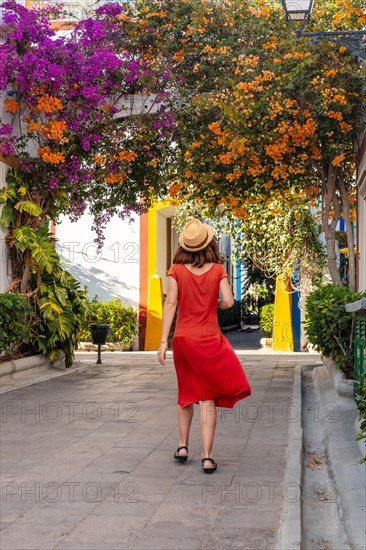 A woman in a red dress walks down a street with a hat on. The street is lined with colorful buildings and trees
