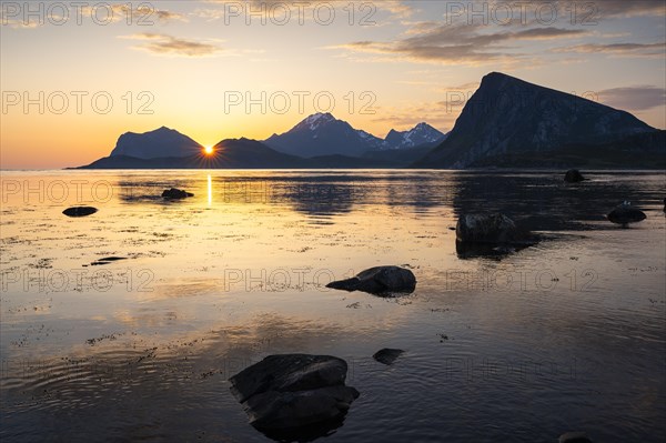Landscape on the Lofoten Islands. View from Flakstadoya to Vestvagoya with the snow-covered mountain Himmeltindan. The mountains are reflected in the sea. The sun is between two mountains, with a sun star. At night at the time of the midnight sun in good weather. Early summer. Lofoten, Norway, Europe