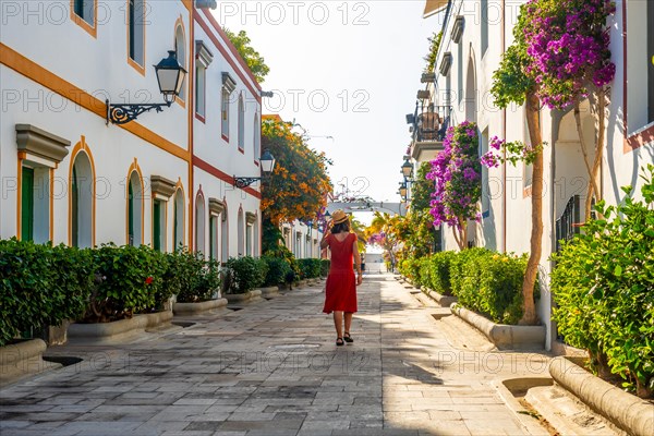 A woman in a red dress walks down a narrow street lined with trees and buildings. The street is lined with potted plants and the woman is wearing a straw hat