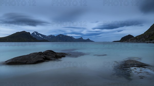 Seascape on the beach at Haukland. View of the mountains of Myrland on Flakstadoya. Dark sky, blurred clouds. Rocks in the foreground. Early summer. Long exposure. Haukland Beach, Haukland, Vestvagoya, Lofoten, Norway, Europe