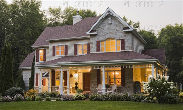 Large illuminated two story beige and tan cut stone home exterior with white and red trim, multiple burgundy asphalt shingles roofs plus Adirondack chairs on veranda supported by wooden columns at dusk in summer, Quebec, Canada, North America