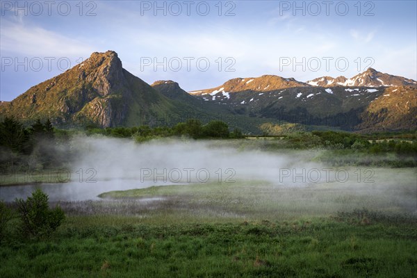 Landscape on the Lofoten Islands. Morning mist rises over a meadow and a stream. Mountains in the background. At night at the time of the midnight sun in good weather, blue sky. Golden hour. Early summer. Vestvagoya, Lofoten, Norway, Europe