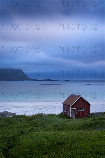 A red wooden hut on the sandy beach of Ramberg (Rambergstranda), sea and mountains. At night at the time of the midnight sun. Clouds in the sky. Early summer. Ramberg, Flakstadoya, Lofoten, Norway, Europe