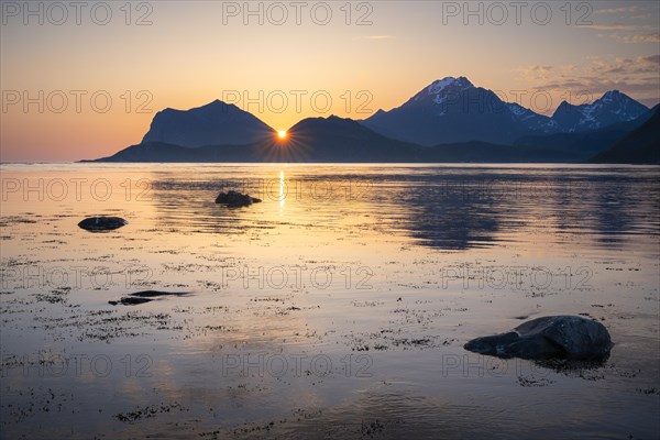 Landscape on the Lofoten Islands. View from Flakstadoya to Vestvagoya with the snow-covered mountain Himmeltindan. The mountains are reflected in the sea. The sun is between two mountains. At night at the time of the midnight sun in good weather. Early summer. Lofoten, Norway, Europe