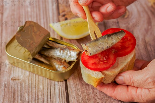 A woman puts a sardine on a slice of bread with tomato, in the background you can see the can of sardines and a lemon on a wooden table