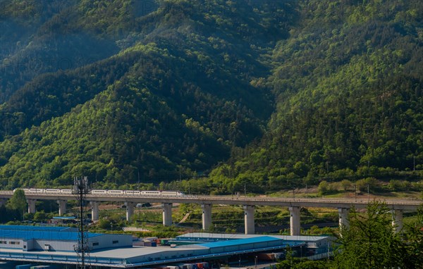 Train bridge spanning over an area with industrial facilities against a backdrop of mountains, in South Korea