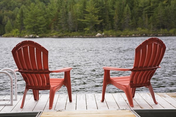 Two bright red plastic Adirondack chairs on wooden floating dock on calm lake with forest of green leaved coniferous and deciduous trees in late summer, Quebec, Canada, North America