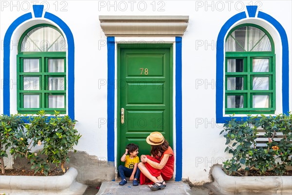 A woman and a child are sitting on the steps of a building with a green door. The woman is wearing a red dress and a straw hat. The scene is peaceful and calm, with the woman