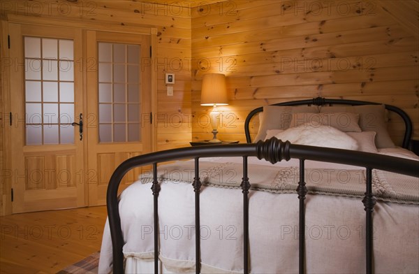 Queen size bed with white bedspread, throw and pillows, tubular cast iron headboard and footboard in master bedroom with pine wood plank walls, french doors and lit night table lamp on upstairs floor inside small log cabin home, Quebec, Canada, North America