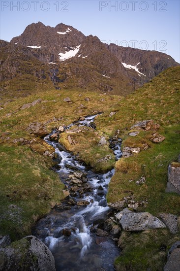 A torrent flows down the Stornappstinden mountain. Long exposure. At night at the time of the midnight sun in good weather, blue sky. Early summer. Flakstadoya, Lofoten, Norway, Europe