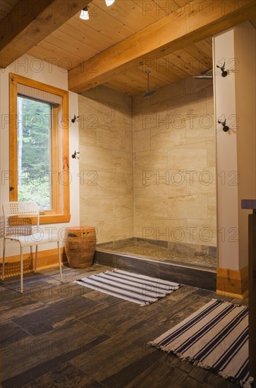 Illuminated beige ceramic shower stall in main bathroom with tan and dark grey ceramic tile floor and blue and white striped rugs on ground floor inside luxurious contemporary timber and milled log cabin home, Quebec, Canada, North America