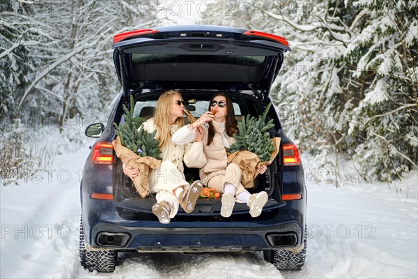 Two joyful women are sitting in the trunk of a car with bouquets of fir branches and drinking champagne