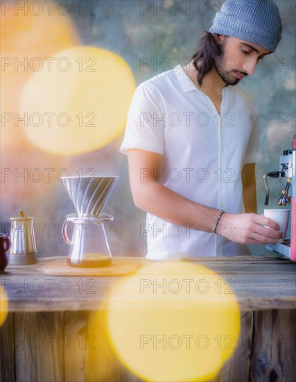 A bearded man in a knit hat prepares a coffee with a machine, soft yellow lights blurred in the foreground, Vertical aspect ratio, AI generated