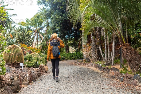 A woman is walking down a path in a garden. She is wearing a yellow jacket and a hat. She is carrying a backpack