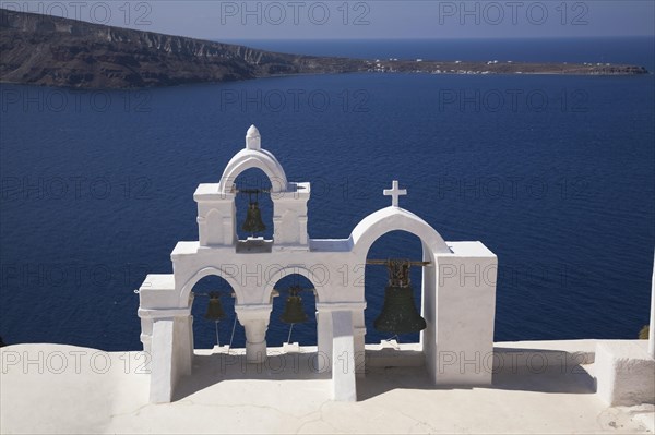 White roughcast cladded arched church bells tower with cross overlooking the Aegean sea, Oia village, Santorini, Greece, Europe