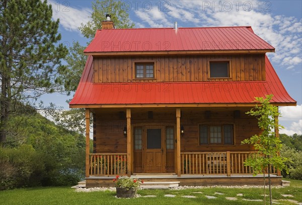 Small two story brown stained log cabin home with veranda and red sheet metal roof and landscaped front yard with deciduous tree and flagstone footpath in summer, Quebec, Canada, North America