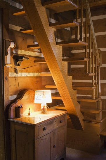 Small antique wooden cabinet with lit lamp underneath a wood staircase made with flat steps supported by a single beam leading to upstairs floor inside white chinked log cabin home, Quebec, Canada, North America