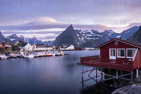 The village of Hamnoy. In the foreground, a typical red wooden house (rorbuer) on wooden stilts. Other houses and boats in a small harbour. Mount Olstinden in the background. At night at the time of the midnight sun in good weather, some clouds in the sky. Early summer. Long exposure. Hamnoy, Moskenesoya, Lofoten, Norway, Europe