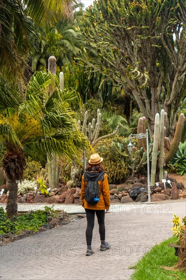 A woman wearing a hat and a backpack walks through a garden. The woman is walking on a path surrounded by trees and plants