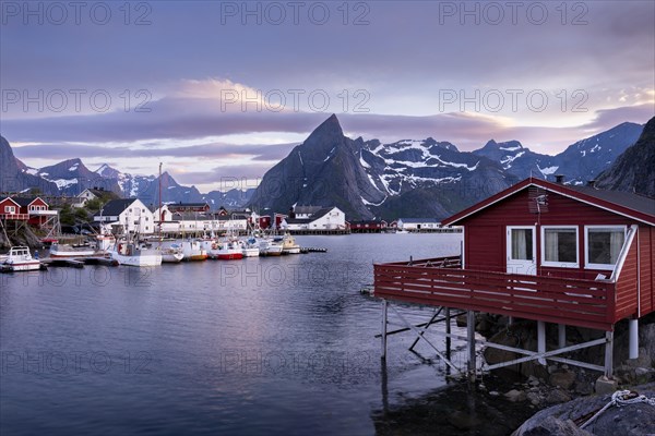 The village of Hamnoy. In the foreground, a typical red wooden house (rorbuer) on wooden stilts. More houses and boats in a small harbour. Mount Olstinden in the background. At night at the time of the midnight sun in good weather, some clouds in the sky. Early summer. Hamnoy, Moskenesoya, Lofoten, Norway, Europe