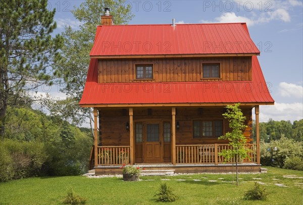 Small two story brown stained log cabin home with veranda and red sheet metal roof and landscaped front yard with deciduous tree and flagstone footpath in summer, Quebec, Canada, North America