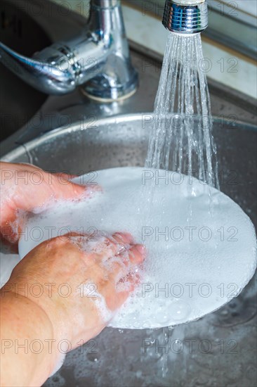 Hands, sink and washing dishes with a person in the kitchen of a home to wash a plate for hygiene. Water, bacteria and soap with an adult cleaning porcelain crockery in a house to clean for housework