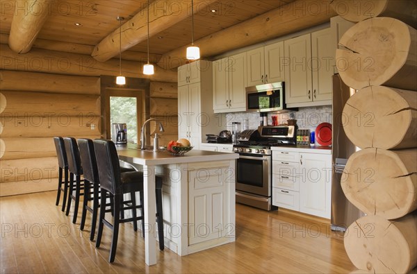 White painted wooden cabinets and island with dark brown wood and leather high back bar stools in kitchen with varnished floor boards inside luxurious contemporary Scandinavian style log cabin home, Quebec, Canada, North America