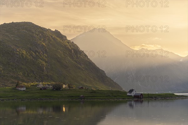 Landscape with sea and mountains on the Lofoten Islands. The fjord Flakstadpollen and some houses belonging to the village of Flakstad. The landscape is reflected in the sea. At night at the time of the midnight sun in good weather, the sun shines in from the side. Flakstadoya, Lofoten, Norway, Europe