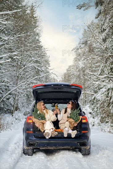 A happy couple is sitting in the trunk of a blue car with bouquets of fir branches, enjoying cozy moments in a snowy forest, clinking glasses with champagne