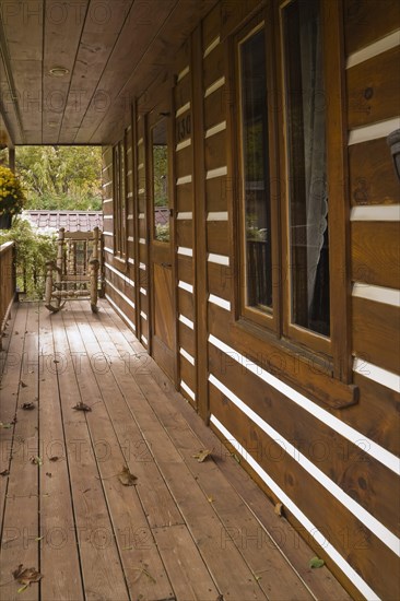 Long old and worn brown stained veranda with pine wood rocking chair on facade of rustic white chinked log cabin home in autumn, Quebec, Canada, North America