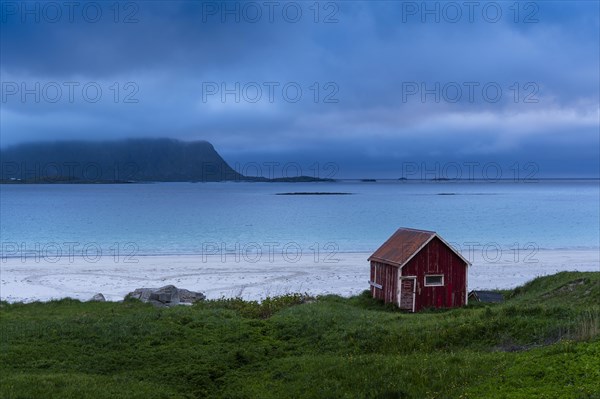 A red wooden hut on the sandy beach of Ramberg (Rambergstranda), sea and mountains. At night at the time of the midnight sun. Clouds in the sky. Early summer. Ramberg, Flakstadoya, Lofoten, Norway, Europe