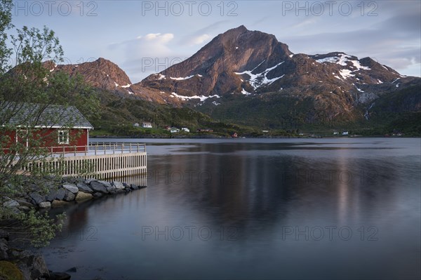 Landscape on the Lofoten Islands. A red wooden house in Kakern on Kakersundet. The mountain Narvtinden on Moskenesoya in the background. The mountain is reflected in the sea. At night at the time of the midnight sun in good weather, a few clouds in the sky. Early summer. Kakern, Flakstadoya, Lofoten, Norway, Europe