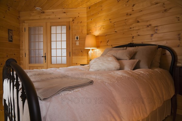 Queen size bed with white bedspread, throw and pillows, tubular cast iron headboard and footboard in master bedroom with pine wood plank walls, french doors and lit night table lamp on upstairs floor inside small log cabin home, Quebec, Canada, North America