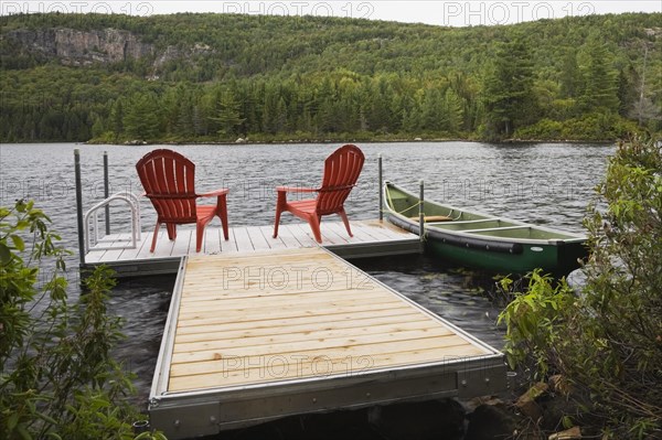 Two bright red plastic Adirondack chairs on floating wooden dock and green rowboat on calm lake with forest of green leaved coniferous and deciduous trees in late summer, Quebec, Canada, North America