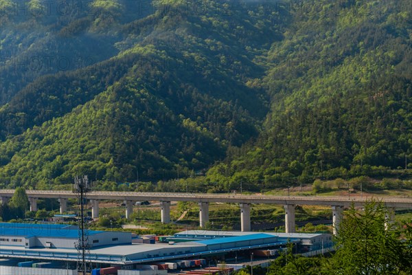 View of a train passing a bridge over an industrial area with mountains in the distance, in South Korea